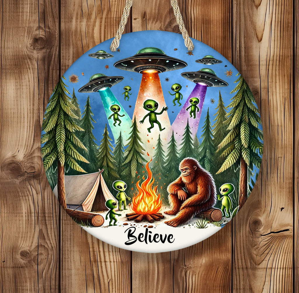 Round 14 inch hardboard round hanging sign featuring Bigfoot sitting by a campfire in a dense pine forest, surrounded by cute green aliens and a glowing tent, with colorful UFOs beaming light down from a star-filled sky — vibrant and whimsical outdoor-themed artwork - Personalized Kreation