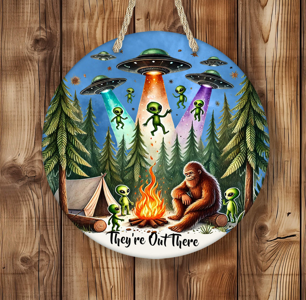 Round 14 inch hardboard round hanging sign featuring Bigfoot sitting by a campfire in a dense pine forest, surrounded by cute green aliens and a glowing tent, with colorful UFOs beaming light down from a star-filled sky — vibrant and whimsical outdoor-themed artwork - Personalized Kreation