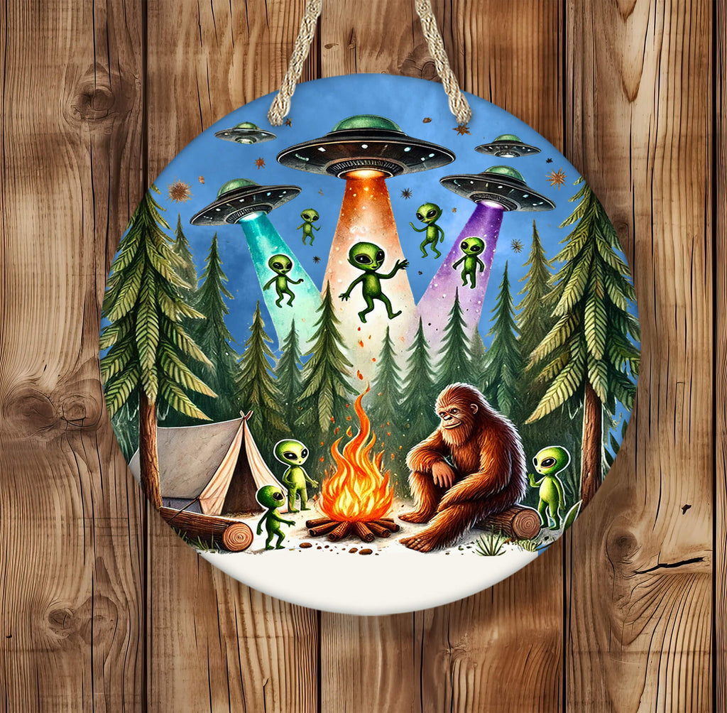 Round 14 inch hardboard round hanging sign featuring Bigfoot sitting by a campfire in a dense pine forest, surrounded by cute green aliens and a glowing tent, with colorful UFOs beaming light down from a star-filled sky — vibrant and whimsical outdoor-themed artwork - Personalized Kreation