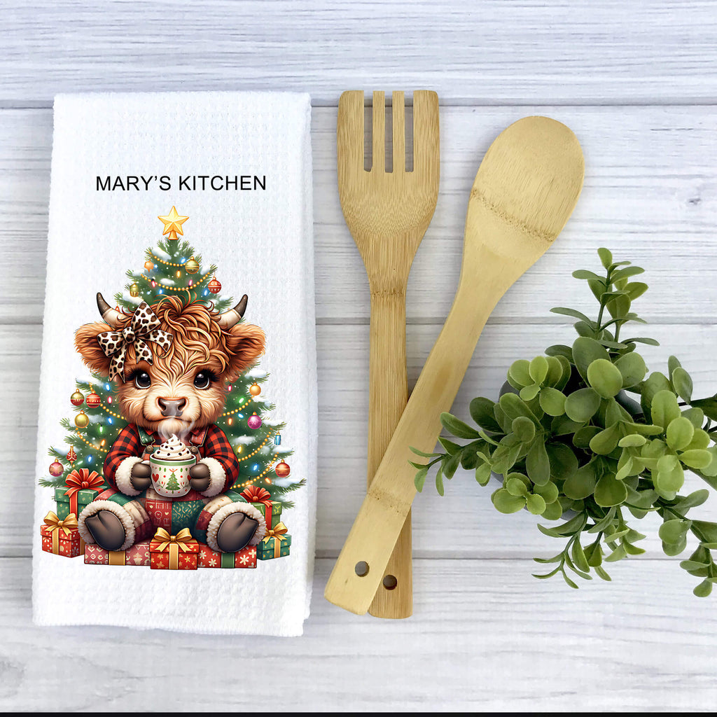 Kitchen towel with a cow under a Christmas tree, next to wooden utensils and a plant.
