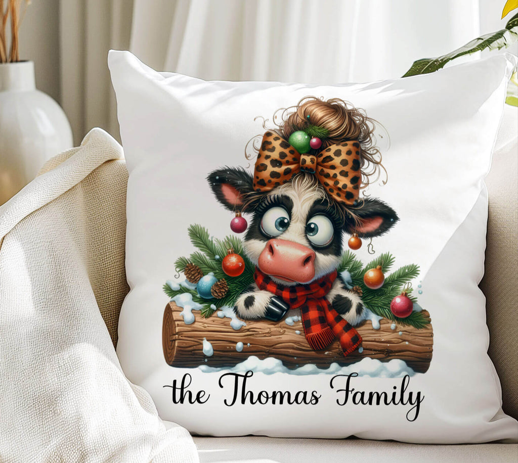 Decorative pillow with a cartoon cow wearing a leopard print hat and plaid scarf, surrounded by Christmas decorations, on a couch.
