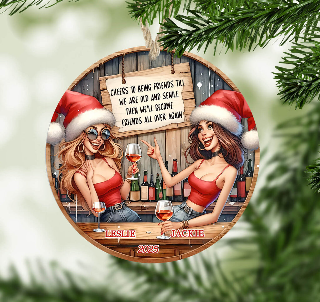 Circular Christmas ornament with two women in Santa hats and red outfits, holding wine glasses, with a wooden sign and bottles in the background.