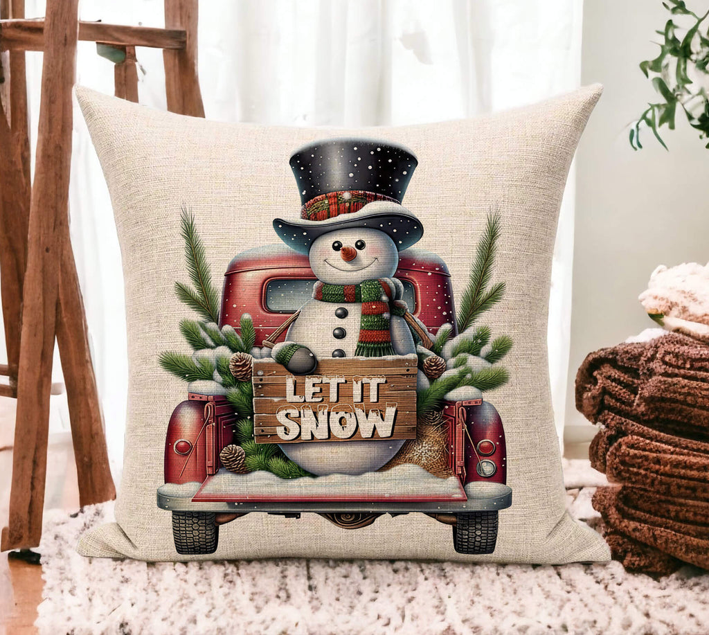 Decorative pillow with a snowman and 'Let It Snow' text on a truck, placed on a surface.