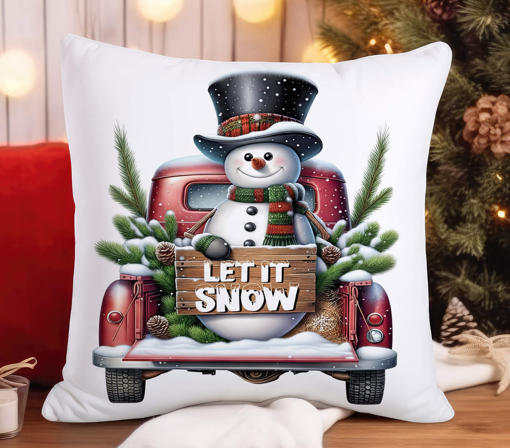 Decorative pillow with a snowman and 'Let It Snow' text on a red truck, placed on a wooden surface with festive decorations.